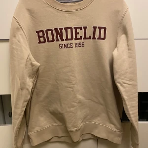 Beige sweatshirt från Bondelid - Säljer en beige sweatshirt från Bondelid med tryck i vinrött framtill. Tröjan har långa ärmar och en rund halsringning. Perfekt för en avslappnad stil.