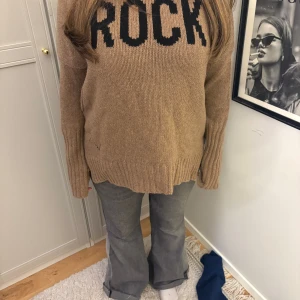 Beige stickad tröja med text - Säljer en beige stickad tröja med texten 'ROCK' i svart på framsidan. Tröjan har långa ärmar och en avslappnad stil. Tröjan är från Zadig.