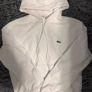 Vit hoodie från Lacoste - Säljer en stilren vit hoodie från Lacoste med klassisk passform. Hoodien har en justerbar huva med dragsko och det ikoniska Lacoste-logotypen på bröstet. Perfekt för en avslappnad look.