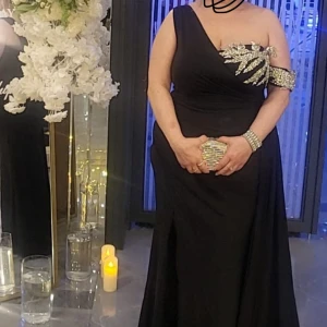 Svart oneshoulder klänning med paljettdetaljer - Elegant svart oneshoulder klänning med glittrande paljettdetaljer på axeln. Klänningen har en lång siluett och en smickrande passform, perfekt för festliga tillfällen.