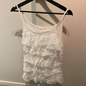 Vit volanglinnetopp - Säljer en söt vit linnetopp med volanger. Toppen har smala axelband och är perfekt för sommardagar. Den är lätt och luftig, vilket gör den idealisk för varma väder.