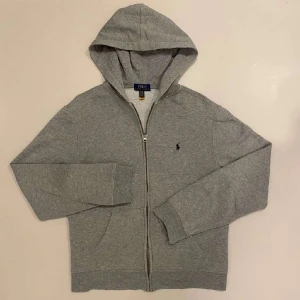Grå hoodie från Ralph Lauren - Säljer en stilren grå hoodie från Ralph Lauren med dragkedja och broderad logga på bröstet. Perfekt för en avslappnad look. Den har en bekväm passform och praktiska fickor framtill. Är small fast passar främst för medium.
