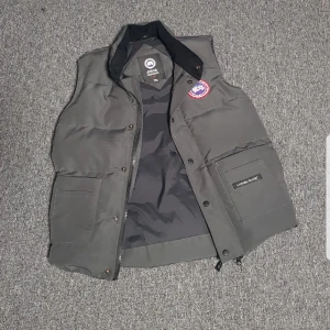  Canada Goose väst - Säljer en svart dunväst från Canada Goose med klassisk design. Västen har en dragkedja och knappar framtill samt en broderad logotyp på bröstet. Perfekt för kyliga dagar.