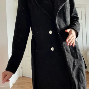 Svart kappa med knappar - Elegant svart kappa med långa ärmar och stilrena knappar framtill. Kappan har en klassisk design med ett bälte i midjan för en figursydd passform. Perfekt för kyligare dagar.