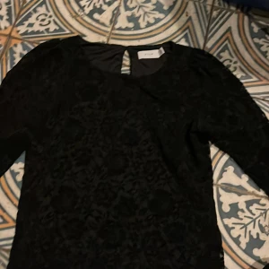 Svart spets tröja från Vila - Elegant svart blus från Aéryne med ett diskret blommönster. Blusen har trekvartsärmar och en rund halsringning med en liten öppning bak. Perfekt för en stilren look.