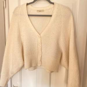 Mysig beige stickad kofta med ballongärm från Gina Tricot med knappar framtill och en v-ringad design. Perfekt för kyliga dagar med sin ribbade struktur och långa ärmar. En stilren och bekväm tröja för alla tillfällen.
