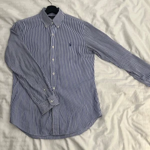 Skjorta från Ralph Lauren - Snygg blå- och vitrandig skjorta från Ralph Lauren. Perfekt för en stilren look.                                               Storlek: M                                                                           Defekt (på bild) knappt synlig på bakre kanten. Skjortan levereras såklart tvättad och strykt