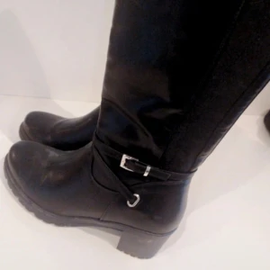 Svarta allvädersstövlar med spänne - Säljer ett par svarta stövlar i skinn med en snygg spänndetalj på sidan. De är i allvädersmaterial vilket gör dem perfekta för både höst och vinter. Stövlarna har en robust sula och en liten klack för extra stil. Perfekta för att hålla fötterna torra och varma under de kallare månaderna.