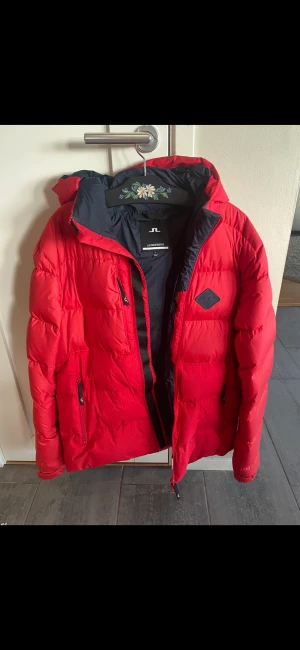 J.Lindberg vinterjacka  - Säljer en röd varm J.Lindberg vinterjacka. Jackan köptes förra hösten för 2700kr. Den är sällan använd och i mycket fint skick. Det enda att anmärka på är en liten lagning gjord i armhålan, som inte syns (Se sista bilden). Hör av er vid frågor!
