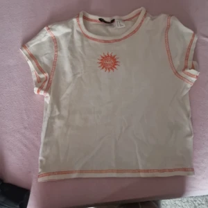 Vit och orange t-shirt från H&M Divided - Säljer en superfin vit t-shirt från H&M Divided i storlek XS. Den har korta ärmar och ett coolt orange tryck med texten 'Social Sunshine' på bröstet. T-shirten har också orange sömmar som ger den en extra touch. Perfekt för sommardagar! 🌞