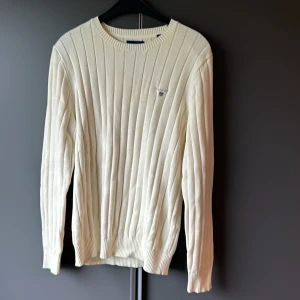 Beige ribbad tröja från GANT - Säljer en stilren och klassisk beige ribbad tröja från GANT. Den är gjord av premium bomull och har en broderad logga på bröstet. Perfekt för både höst och vår, med långa ärmar och en bekväm passform. Passar lika bra till vardags som till lite finare tillfällen.