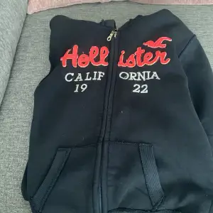 Säljer en svart hoodie från Hollister med dragkedja och tryck på framsidan. Den har texten 'Hollister California 1922' i rött och vitt. Perfekt för en avslappnad stil och passar bra till höst och vinter. Supermjuk och bekväm! Använd två gånger!