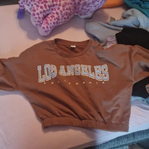 Brun croppad tröja från Lindex - Säljer en supercool brun croppad tröja från Lindex med texten 'Los Angeles California' på framsidan. Perfekt för en avslappnad stil och passar bra till jeans eller leggings. Tröjan är i bra skick och redo för nya äventyr!