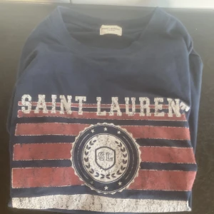 Mörkblå t-shirt från Saint Laurent - Säljer en snygg mörkblå t-shirt från Saint Laurent med ett coolt tryck i rött och vitt. Perfekt för en avslappnad stil. T-shirten har en normal passform och är gjord i mjuk bomull. Passar perfekt till både jeans och shorts! Nypris: runt 5000. Mitt pris: 1850