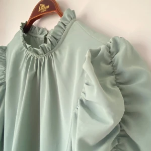 Ljusgrön blus från ONLY - Superfin ljusgrön blus från ONLY med volangdetaljer vid hals och ärmar. Perfekt för vår och sommar! Blusen har en lös passform och korta ärmar med puffiga volanger. Den är gjord i ett mjukt och bekvämt material, perfekt för både vardag och fest.