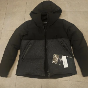 Svart dunjacka från Sail Racing - Säljer en svart dunjacka från Sail Racing ,i storlek L. Jackan är i nytt skick med etiketter kvar. Den har en luva och dragkedja framtill. Perfekt för kalla vinterdagar med sitt 700 fill power dun. Super varm och fin. Jackan är helt ny ,bara provad en gång, aldrig använd. Orginalpris 5000kr men säljer för 1800kr.
