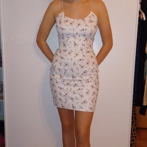 Blommig kortklänning med smala axelband - Söt och somrig kortklänning med blommigt mönster. Klänningen har smala axelband och en tight passform som framhäver figuren. Perfekt för varma sommardagar eller en avslappnad fest. Materialet känns lätt och bekvämt mot huden.