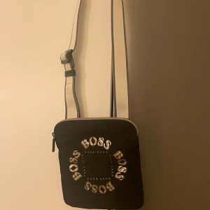 Svart axelväska från Boss - Snygg svart axelväska från Boss med justerbar rem och dragkedja. Väskan har en stilren design med Boss-loggan i silverfärgade bokstäver på framsidan.