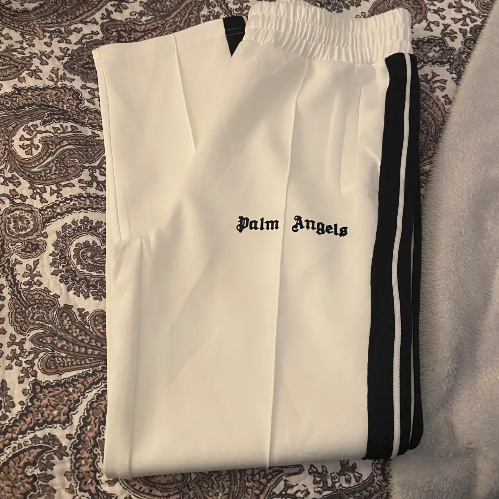 Säljer ett par vita byxor från Palm Angels i mycket bra skick. De har en elastisk midja och svarta ränder längs sidorna. Perfekta för en avslappnad och stilren look. Passar både till vardags och festliga tillfällen.. Jeans & Byxor.