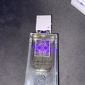 Parfym från Lattafa - Säljer en elegant parfym från Lattafa. Flaskan är genomskinlig med ett stilrent, fyrkantigt lock i silver. På framsidan finns ett dekorativt mönster i lila och silver. Perfekt för den som vill ha en unik doftupplevelse. Flaskan har en modern och lyxig design.