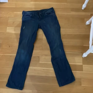 Lågmidjade Blå jeans från Wrangler - W27 L32 Säljer ett par klassiska blå jeans från Wrangler. De har en snygg, sliten look och är i bra skick. Perfekta för vardagsbruk med en straight passform. Märkesloggan syns tydligt på bakfickan. Passar till alla tillfällen, både avslappnade och lite mer uppklädda. 
