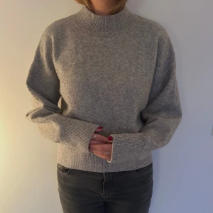 Grå stickad tröja - Mysig grå stickad tröja med hög krage och långa ärmar. Perfekt för kyliga dagar och passar både till vardags och lite finare tillfällen. Tröjan har en ribbad kant vid ärmslut och nederkant för extra komfort.