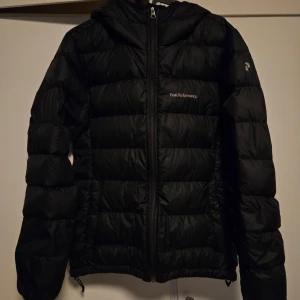 Svart dunjacka från Peak Performance - Säljer en svart dunjacka från Peak Performance i mycket bra skick. Jackan har en dragkedja framtill och en mysig huva. Perfekt för kalla vinterdagar och höstpromenader. Den är både stilren och funktionell, vilket gör den till ett måste i garderoben!