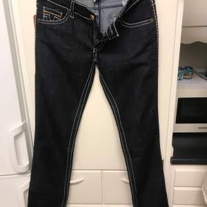 Säljer ett par snygga mörkblå jeans från True Religion i storlek 30. De har vita kontrastsömmar och klassiska femficksdesign. Jeansen är tillverkade i USA av 100% bomull och har en bekväm passform. Perfekta för både vardag och fest!