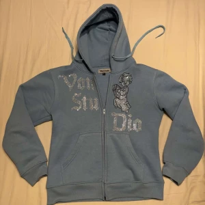 Blå hoodie från Vonstudio - Säljer en snygg blå hoodie från Vonstudio i storlek S. Den har en dragkedja framtill och ett coolt tryck med text och en figur på framsidan. Perfekt för en avslappnad stil och passar bra till både jeans och joggers. Huvan har dragsnören för justering.