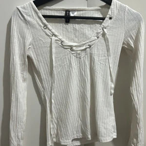 Vit ribbad tröja från H&M Divided - Säljer en snygg vit ribbad tröja från H&M Divided i storlek XS. Tröjan har långa ärmar och en cool snörning framtill som ger den en unik touch. Perfekt för både vardag och fest! 🖤