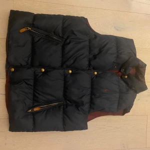 Svart dunväst med broderad logga - Säljer en snygg svart dunväst med guldknappar och dragkedja. Västen har en broderad logga på bröstet och är perfekt för höst och vinter. Den är i mycket bra skick och har en normal passform. Perfekt för att hålla värmen med stil!