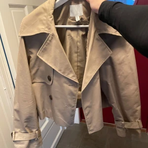 Beige trenchcoat - Säljer en snygg beige trenchcoat i nyskick. Jackan har en klassisk design med stora knappar och bälte vid ärmsluten. Perfekt för höst och vår, och ger en stilren look. Passar till både vardag och fest!