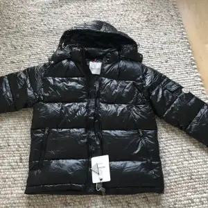 Säljer en snygg svart dunjacka från Moncler i nytt skick. Jackan har en glansig finish och är perfekt för kalla vinterdagar. Den har en dragkedja framtill och en huva för extra värme. Moncler-loggan syns på ärmen, vilket ger en exklusiv touch. Perfekt för dig som vill hålla stilen i vinter! 🖤