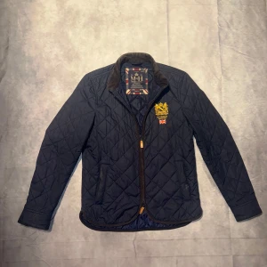 Mörkblå quiltad jacka från Morris - Säljer en snygg mörkblå quiltad jacka från Morris. Den har en broderad logga på bröstet och en dragkedja framtill. Perfekt för höst och vår med sin stilrena design och bekväma passform. Jackan är långärmad och har en klassisk krage. Passar både till vardags och lite finare tillfällen. Storlek Xsmall sitter som small.