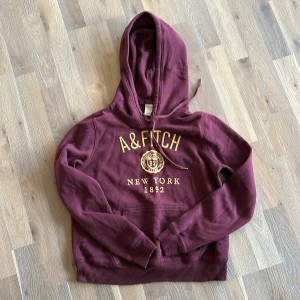 Vinröd hoodie från A&Fitch - Säljer en snygg vinröd hoodie från A&Fitch med gul text och emblem på framsidan. Den har en klassisk känguruficka och en justerbar huva. Perfekt för höst och vinter, supermysig och stilren! 🧡