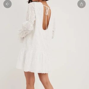 Vit klänning med öppen rygg - Superfin vit klänning med öppen rygg och knytning. Den har långa ärmar med volangdetaljer och ett vackert broderat mönster. Perfekt för studenten. Klänningen är kort och har en luftig passform. ENDAST ANVÄND 1 GGN. 