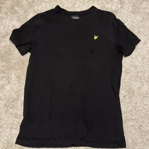 Svart t-shirt från Lyle & Scott  - Säljer en svart t-shirt från Lyle & Scott i mycket bra skick. Den har en broderad gul logga på bröstet. Perfekt för vardagsbruk eller en avslappnad stil. Passar ungdomar i åldern 14-15 år.