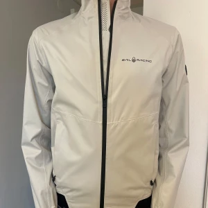 Vit vindjacka från Sail Racing - Säljer en stilren vit vindjacka från Sail Racing i storlek XS. Jackan har en hög krage och dragkedja framtill. Perfekt för blåsiga dagar och har en sportig look. Den är i mycket bra skick och passar både till vardags och träning. 