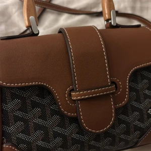 Brun och mönstrad handväska - Snygg brun handväska med unikt mönster och trähandtag. Väskan har en stilren design med vita sömmar och en praktisk axelrem. Perfekt för både vardag och fest! Det långa bandet är avtagbart. Priset går att diskutera vid snabb affär. 