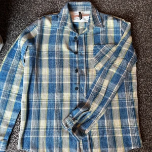 Rutig skjorta från Nudie Jeans Co - Säljer en helt ny blå och gul rutig skjorta från Nudie Jeans. Använd en gång. Inga defekter. Är ganska tjockt material så är lite varmare än en vanlig skjorta. 