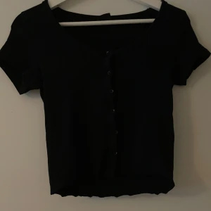 Svart kortärmad topp med knappar - Säljer en stilren svart topp med korta ärmar och knappar framtill. Perfekt för både vardag. Toppen har en enkel och klassisk design som passar till det mesta. Den är i bra skick och redo för en ny ägare!