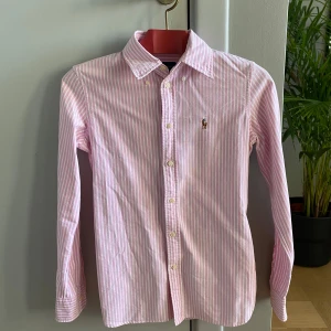 Rosa och vit randig skjorta från Ralph Lauren - Säljer en snygg rosa och vit randig skjorta från Ralph Lauren i storlek XS. Skjortan har långa ärmar och klassisk krage med knappar hela vägen ner. Perfekt för både vardag och fest! 💖