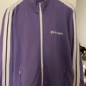 Lila track jacket från Palm Angels - Säljer en snygg lila track jacket från Palm Angels. Den har vita och svarta ränder längs ärmarna och en dragkedja framtill. Perfekt för en sportig look eller chill vardagsstil. Jackan är i bra skick och redo för nya äventyr!