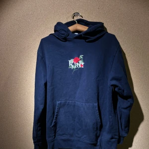 Mörkblå Kith hoodie med rosdetalj - Säljer en snygg mörkblå Kith hoodie med en cool rosdetalj på bröstet. Den har en stor ficka fram och text på huvan. Perfekt för höst och vinter, supermysig och stilren. Passar både till vardags och chillkvällar. 🥶🌹
