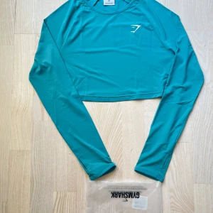 Turkos långärmad topp från Gymshark - Säljer en snygg turkos långärmad topp från Gymshark. Perfekt för träning med sin croppade design och bekväma passform. Materialet är mjukt och stretchigt, vilket gör den idealisk för både gymmet och vardagsaktiviteter. Toppen är i nyskick och kommer i originalförpackning.