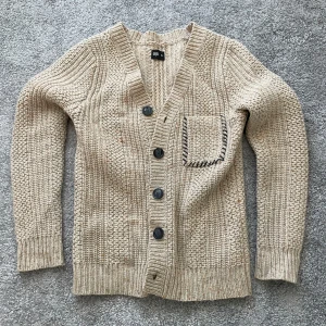 Beige stickad kofta med fickdetalj - Säljer en mysig beige stickad kofta med snygga knappar och en unik ficka med mörkare kantdetalj. Koftan är i nyskick, clean och enkel att stylea.