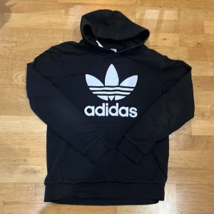 Svart hoodie från Adidas - Säljer en svart hoodie från Adidas med den klassiska loggan i vitt på framsidan. Den är i bra skick och perfekt för både vardag och träning. Hoodien har långa ärmar och en bekväm passform. Mycket fint skick!