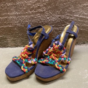 Färgglada kilklackar med flätade detaljer - Säljer ett par unika kilklackar med blå bas och färgglada flätade remmar i rött, gult, grönt och lila. Skorna har en bekväm korksula och justerbara remmar för perfekt passform. Perfekta för sommarens fester eller en dag på stan!