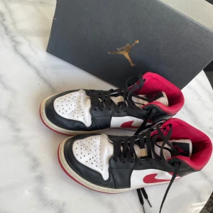 Nike Air Jordan 1 Mid i svart, vitt och rött - Säljer ett par Nike Air Jordan 1 Mid i klassiska färger: svart, vitt och rött. Skorna har snörning och är i bra skick med en cool design som passar perfekt för både vardag och fest. De är bekväma och stilrena, ett måste för sneakerheads!
