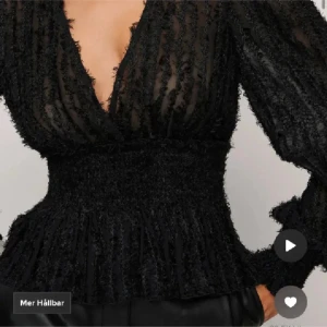 Svart blus med volangdetaljer - Snygg svart blus med volangdetaljer och djup v-ringning. Den har långa ärmar och en smockad midja som ger en fin siluett. Perfekt för en kväll ute eller en festlig tillställning som nu till jul o h nyår.